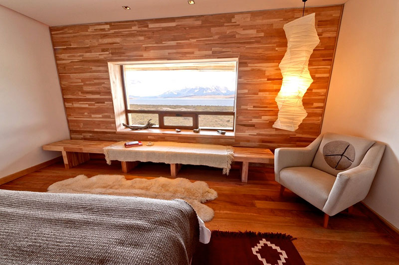 A imagem mostra um quarto de hotel moderno com uma vista deslumbrante da paisagem patagônica. O quarto possui uma grande janela que oferece uma vista panorâmica da natureza. A decoração é minimalista e elegante, com uma paleta de cores que inclui tons de marrom, bege e branco. Os móveis incluem uma cama coberta com edredons de tricô, uma poltrona branca com um travesseiro circular, e uma mesa de centro de madeira. O chão é adornado com tapetes de pele e um tapete com padrões geométricos. A parede de madeira e a iluminação suave contribuem para a atmosfera acolhedora e serena.