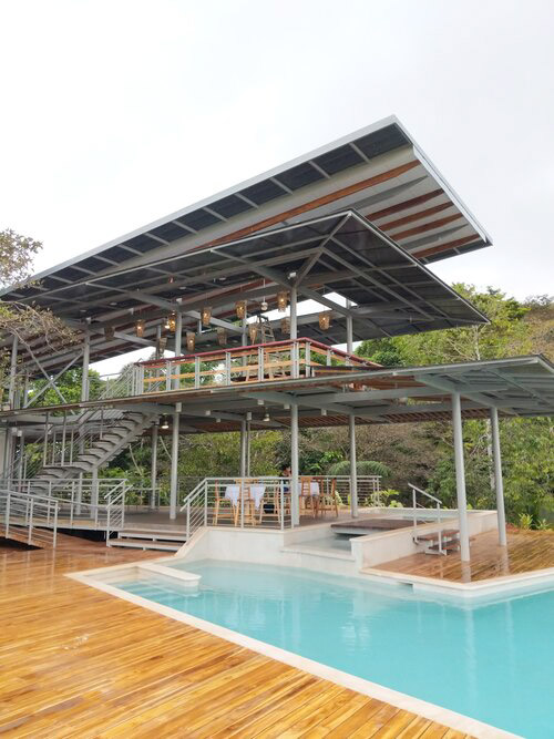 A imagem mostra uma área externa moderna do Cielo Lodge na Costa Rica, caracterizada por uma piscina azul clara cercada por um deck de madeira. A área de estar ao ar livre possui móveis de madeira e metal, com um telhado de metal corrugado proporcionando sombra. A decoração é minimalista e moderna, com elementos naturais e vegetação ao fundo.