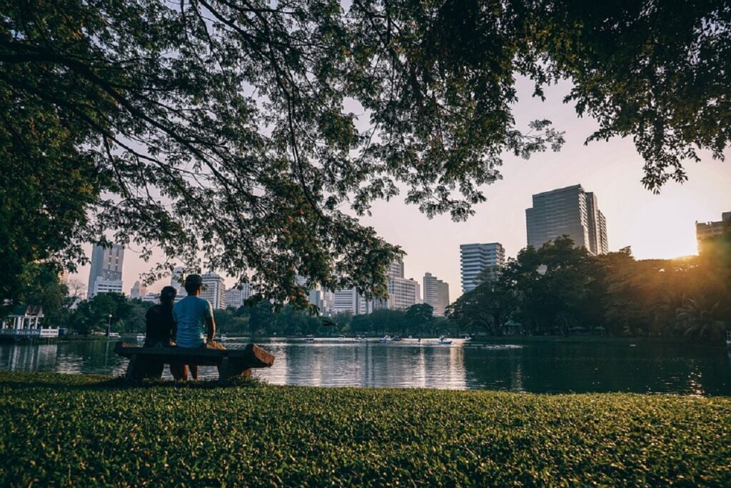 Lago de um parque com um gramado em frente e um casal sentado em um banco olhando a cidade e o pôr do sol ao fundo
