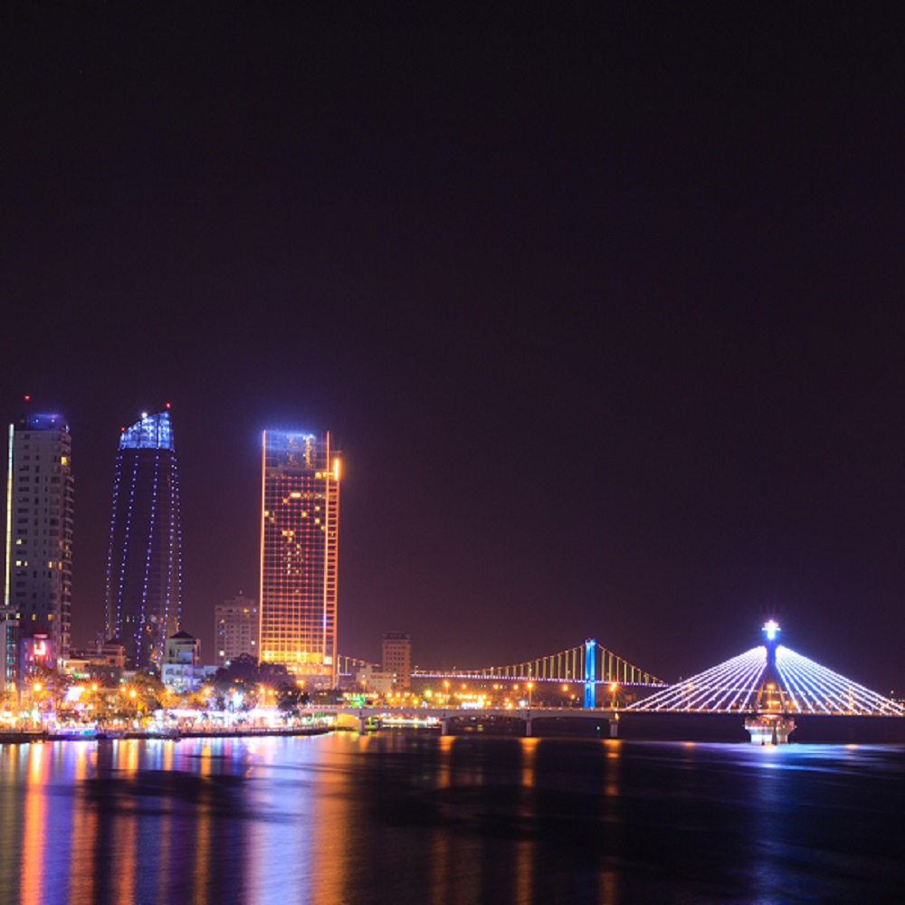 Imagem noturna de Seul, com o Rio Han em destaque e ao fundo os arranha céus iluminados, e a bela ponte estaiada também iluminada em tom azul.
