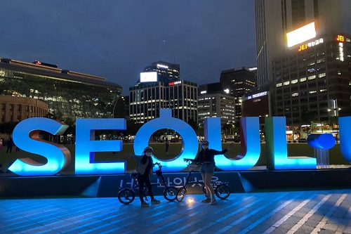2 pessoas de bicicleta na frente do letreiro de cor azul com a palavra Seul. É noite.