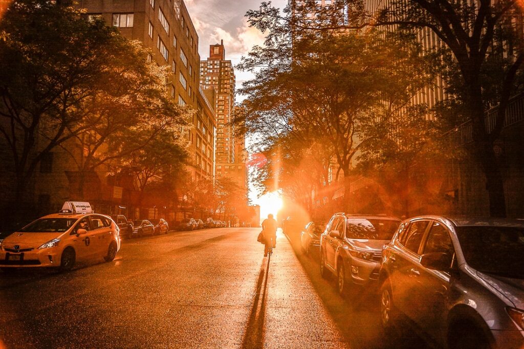 Rua de Nova York, com o sol se ponto no final da rua, e por isso da imagem amarelada. Árvores com folhas de ambos os lados. Carros estacinados de ambos os lados, sendo o da direita um clássico táxi amarelo. Uma pessoa bem longe caminhando de costas, como se fosse em direção ao sol.