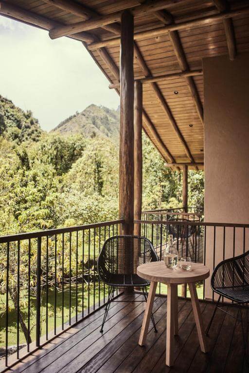 A imagem mostra uma varanda serena no Hotel Andenia Boutique Hotel, no Peru. A varanda possui uma mesa de madeira redonda com duas cadeiras de vime preto, criando um ambiente acolhedor. A varanda é cercada por um corrimão de metal e oferece uma vista deslumbrante de uma paisagem verdejante e montanhosas. O design é moderno e minimalista, com materiais como madeira e metal predominando.