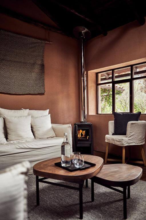 A imagem mostra uma sala de estar moderna no Hotel Andenia Boutique Hotel, no Peru. A sala apresenta uma paleta de cores quentes com tons terrosos nas paredes e elementos de madeira. O móvel principal é um sofá branco com várias almofadas, localizado ao lado de uma lareira. Na frente do sofá, há uma mesa de centro de madeira com duas taças de vinho e uma garrafa. Ao lado da mesa, há uma cadeira de madeira com almofadas. A janela oferece uma vista para a vegetação externa, e há um tapete de cor clara no chão. A decoração é minimalista e elegante, com um toque de conforto.