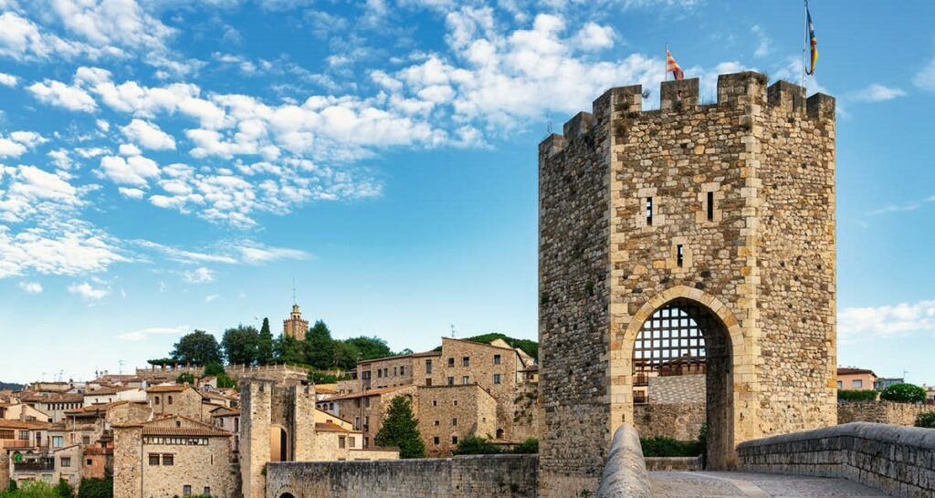 Ponte medieval, construída com pedras, no vilarejo de Besalu (Espanha). Torre de Pedra com portão de ferro semi aberto. Uma bandeira da Espanha estirada do topo da torre que está no centro da ponte. Ao fundo, mais edifícios do vilarejo, que faz parte da Catalunha.