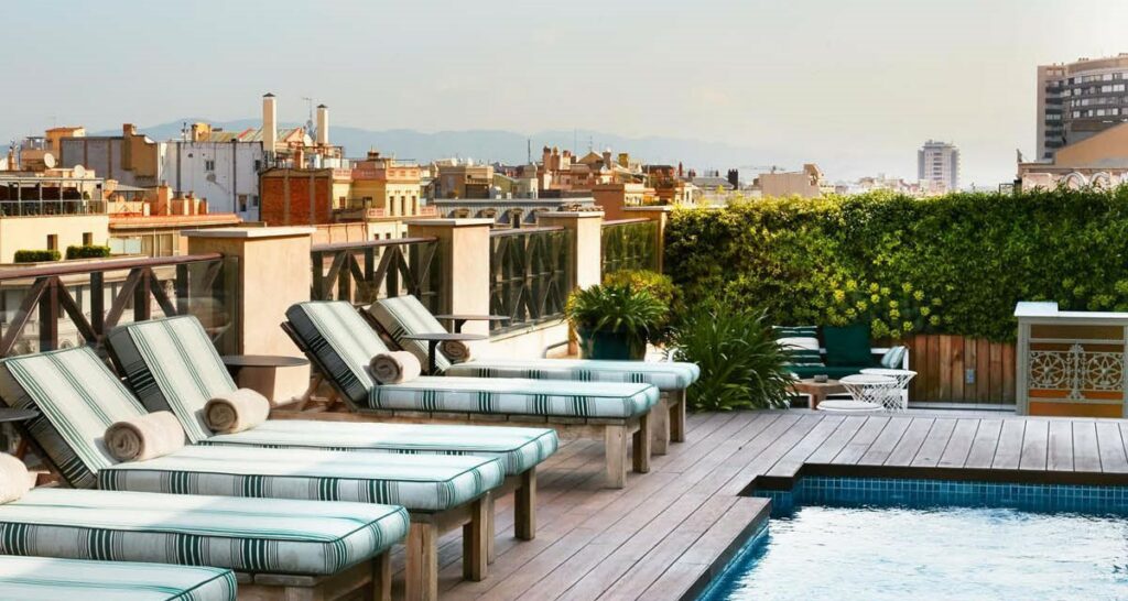 Visão panorâmica do topo dos edifícios em Barcelona (Espanha) a partir do rooftop de hotel Cotton. Várias espreguiçadeiras ao redor de uma piscina no espaço do hotel.