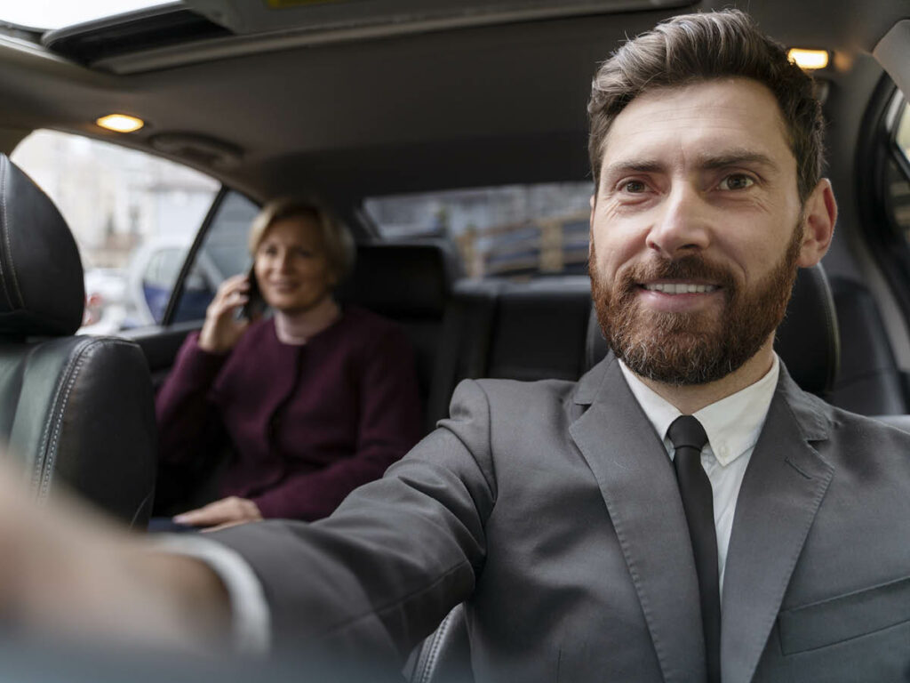 Motorista em serviço de transfer, utilizando terno, sorrindo para a câmera. Passageira ao fundo. 