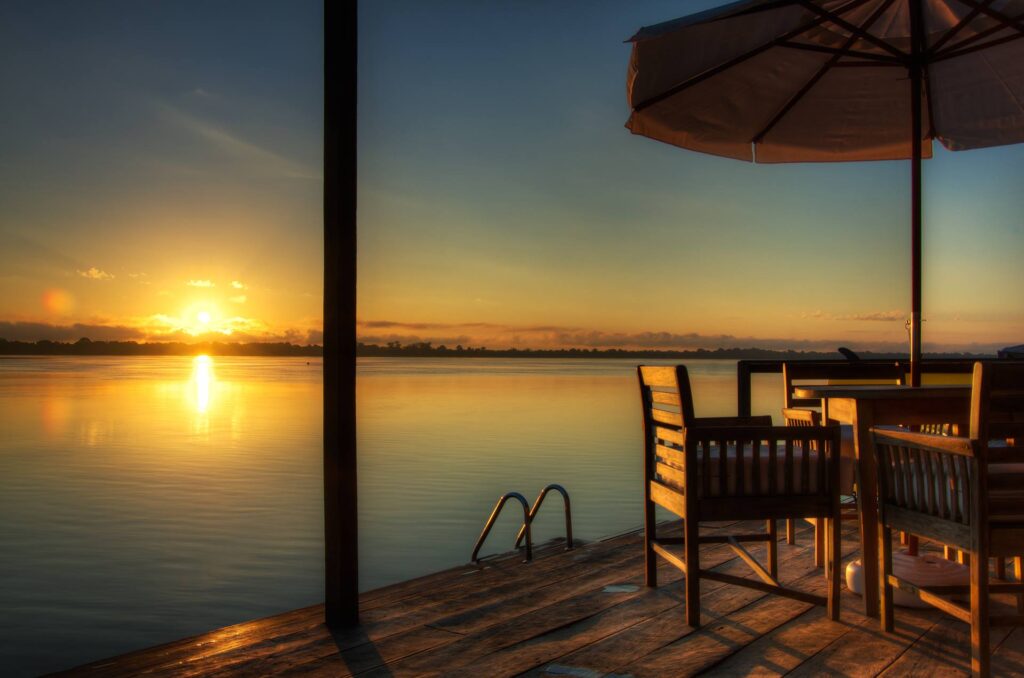 A imagem captura um pátio sereno ao pôr do sol no Lodge do Instituto Ana Vilhanas, na Amazônia brasileira. O pátio possui móveis de madeira, incluindo cadeiras e uma mesa, com um guarda-sol proporcionando sombra. A vista oferece um horizonte tranquilo com o sol se pondo no horizonte, refletindo nas águas calmas do rio. O ambiente é cercado por uma atmosfera natural e pacífica.