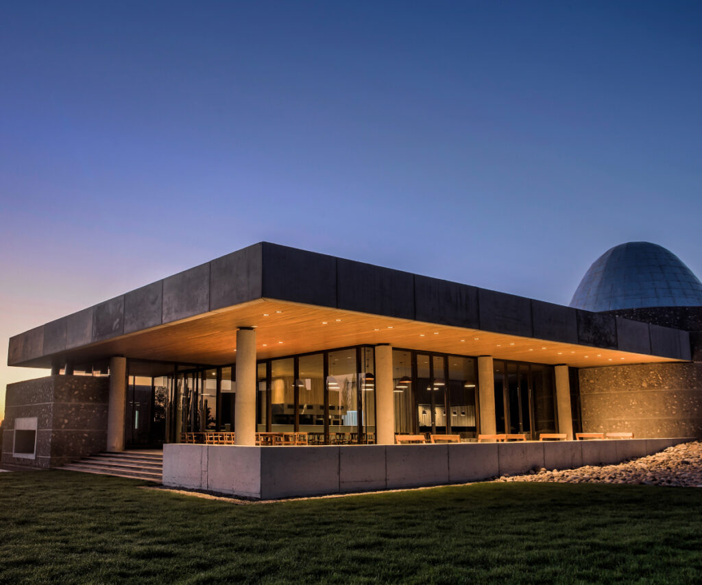 A imagem mostra o exterior de um restaurante moderno chamado Piedra Infnita Cocina I, localizado no Valle de Uco. O edifício apresenta uma arquitetura contemporânea com uma combinação de concreto e vidro. A fachada é iluminada por luzes quentes que destacam a estrutura e criam uma atmosfera acolhedora. O restaurante é cercado por um gramado bem cuidado e pedras decorativas.