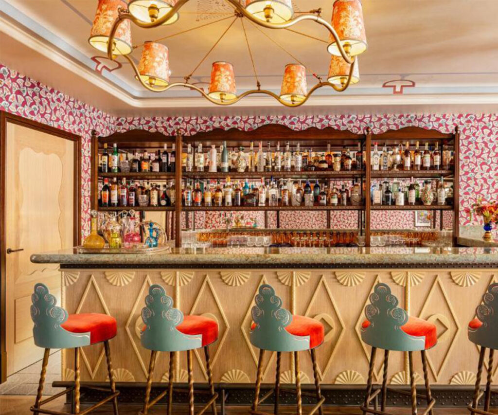 bar do hotel le grand mazarin. Bar estiloso com paredes decoradas com padrões rosa e branco. A iluminação é proporcionada por um lustre com cúpulas laranja e amarelas. O balcão de granito abriga várias garrafas de bebidas e copos. Atrás do balcão, prateleiras cheias de garrafas de bebidas alcoólicas. Cadeiras com assentos vermelhos e encostos verdes estão arrumadas na frente do balcão. A atmosfera é moderna e convidativa, ideal para um ambiente de bar sofisticado.