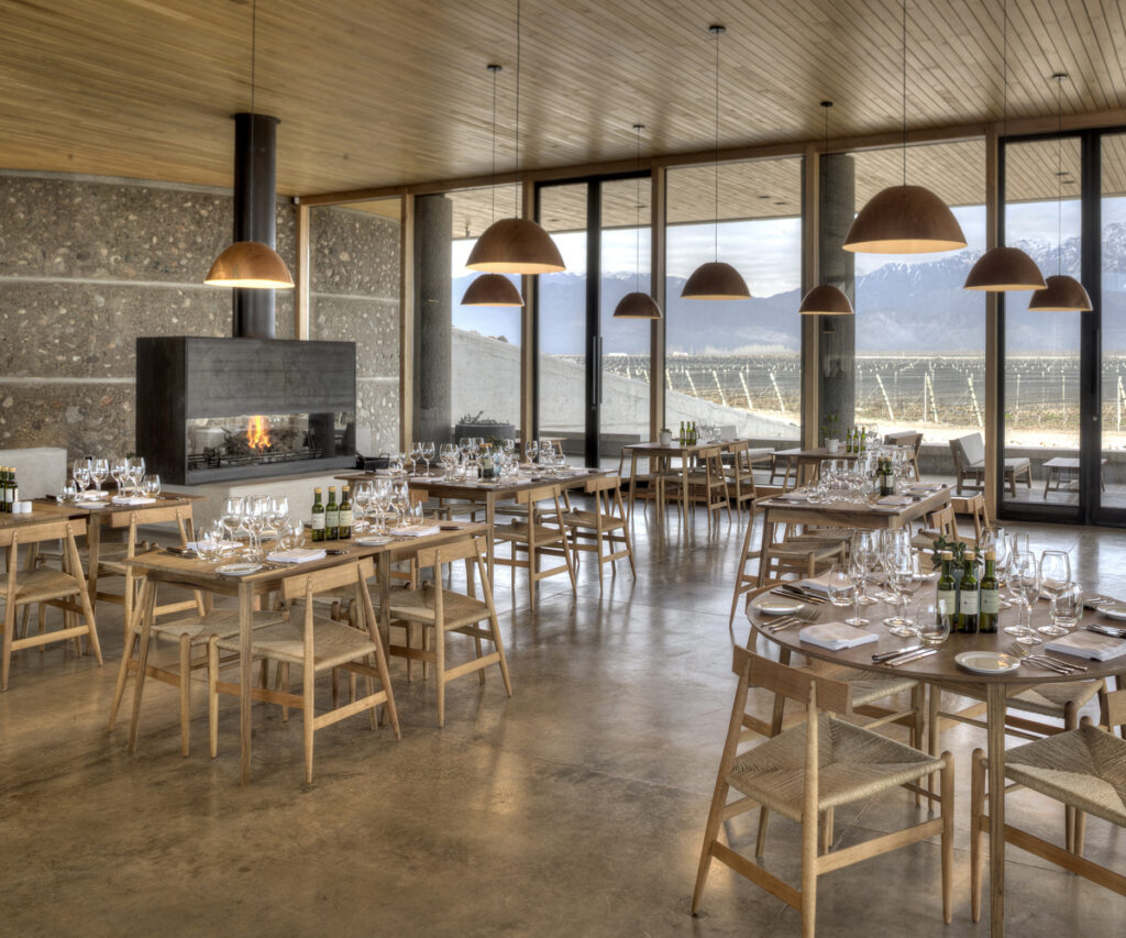 O restaurante Piedra Infinita Cocina, localizado dentro da vinícola Zuccardi, apresenta um design interior moderno com uma paleta de cores neutras. A decoração inclui mesas de madeira com cadeiras de vime, iluminação pendente em formato de cúpula e uma lareira central. As grandes janelas oferecem uma vista deslumbrante dos vinhedos e montanhas ao fundo.