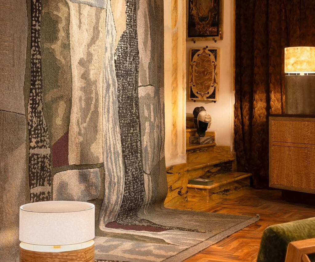 espaço do maison objet - sala com escada, tapete e luminárias. Um quarto de hotel elegantemente decorado com um tapete multicolorido e texturizado cobrindo o chão. O tapete apresenta tons de cinza, verde e marrom, com padrões abstratos. Ao fundo, há uma cama com lençóis brancos e uma cabeceira dourada, adornada com duas pinturas emolduradas nas paredes. A iluminação suave é proporcionada por duas luminárias, uma em cada lado do quarto, criando uma atmosfera quente e acolhedora. A composição é equilibrada, com elementos de design que evocam um senso de conforto e sofisticação.