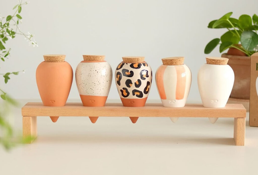 Cinco vasos decorativos de cerâmica, expostos no Maison & Objet Paris, estão arrumados em uma prateleira de madeira. Os vasos apresentam cores e padrões variados: um é laranja terroso, outro tem um acabamento branco com pontos pretos, o terceiro possui um padrão de leopardo, o quarto mistura tons de laranja e branco, e o último é completamente branco. Todos têm tampas de cortiça e estão posicionados em uma linha horizontal, com plantas verdes em vasos ao fundo, criando uma atmosfera acolhedora e esteticamente agradável. A composição destaca a diversidade de estilos e a harmonia entre elementos naturais e feitos pelo homem.