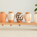 Cinco vasos decorativos de cerâmica, expostos no Maison & Objet Paris, estão arrumados em uma prateleira de madeira. Os vasos apresentam cores e padrões variados: um é laranja terroso, outro tem um acabamento branco com pontos pretos, o terceiro possui um padrão de leopardo, o quarto mistura tons de laranja e branco, e o último é completamente branco. Todos têm tampas de cortiça e estão posicionados em uma linha horizontal, com plantas verdes em vasos ao fundo, criando uma atmosfera acolhedora e esteticamente agradável. A composição destaca a diversidade de estilos e a harmonia entre elementos naturais e feitos pelo homem.