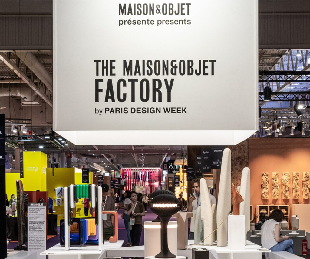 A imagem mostra uma exposição na feira Maison&Objet em Paris, apresentando o 'The Maison&Objet Factory by Paris Design Week'. O ambiente é um grande salão de exposições com várias peças de design moderno, incluindo luminárias, móveis e decorações. A paleta de cores predominante inclui branco, preto e tons vibrantes como amarelo e verde. Materiais como metal, vidro e cerâmica são visíveis. O estilo de decoração é moderno, com um toque industrial. O ambiente parece ser uma feira de design, com várias pessoas interagindo com os produtos expostos.