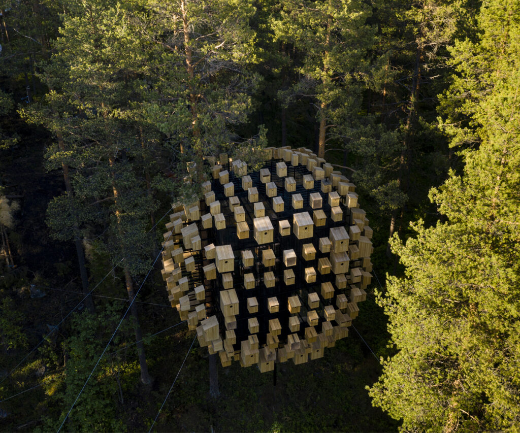 A imagem mostra a Biosfera do Tree Hotel, um hotel de design único na Suécia. A estrutura é uma esfera suspensa entre árvores, composta por múltiplos módulos retangulares. A biosfera está localizada em meio a uma floresta densa, cercada por árvores verdes exuberantes. A estrutura é sustentada por cabos e parece integrar-se harmoniosamente com o ambiente natural.