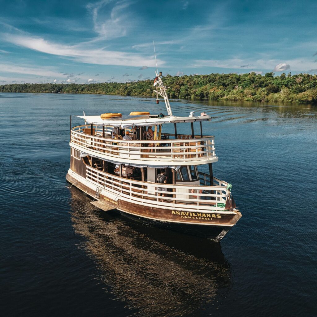 Viagem para Amazônia. A imagem mostra um barco do Ana Vilhanas Lodge navegando em um rio na Amazônia. O barco tem dois níveis com corrimãos de madeira e um convés superior aberto. O barco está cercado por uma paisagem verde exuberante e um céu azul claro.