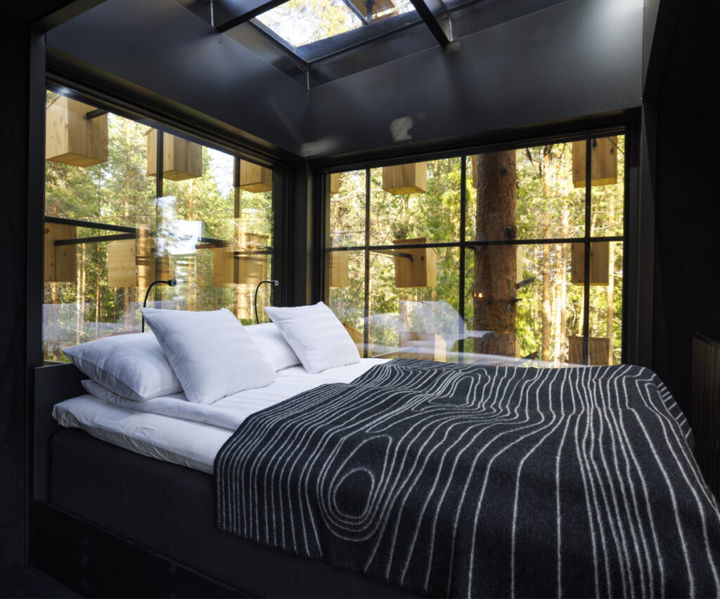 O quarto apresenta uma cama king-size com lençóis brancos e um cobertor cinza com padrão geométrico. As paredes e o teto são escuros, contrastando com a luz natural que entra pelas grandes janelas de vidro. A vista oferece uma paisagem serena de árvores e vegetação, proporcionando uma sensação de tranquilidade e conexão com a natureza. Estilo Moderno para o turismo do sono