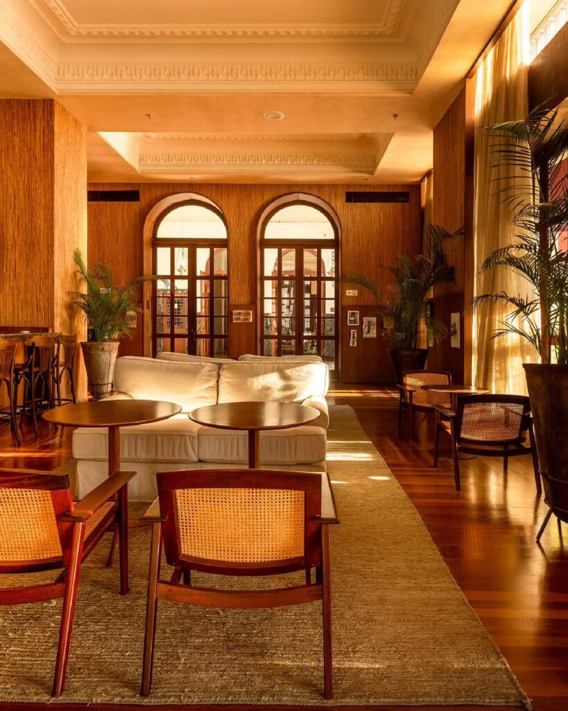 Viagem naBahia: Sala de estar luxuosa no Hotel Fasano Salvador, na Bahia, com móveis de madeira e tons quentes. A sala apresenta um sofá branco, cadeiras de vime e mesas de centro circulares. Arcos e portas de vidro com molduras de madeira conduzem a um pátio externo. Plantas em vasos e cortinas longas complementam a decoração. A iluminação suave cria uma atmosfera aconchegante e sofisticada, destacando a elegância do design interior.