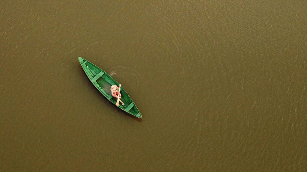 A imagem mostra uma pessoa em um barco verde navegando em um rio com água marrom. O barco é pequeno e parece ser feito de madeira ou material similar. A pessoa está sentada no barco, usando uma camisa rosa. A cena é tranquila e parece ser uma área natural, na Amazônia.