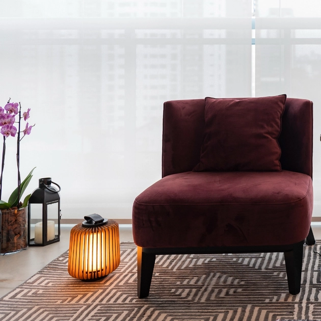A imagem apresenta uma sala de estar moderna com uma cadeira de veludo vermelho como ponto focal. A cadeira está posicionada sobre um tapete com padrão geométrico. Ao lado da cadeira, há uma luminária de mesa de madeira com tecido imbuia steche e uma lanterna preta. Uma planta com flores roxas adiciona um toque de natureza ao ambiente. A janela ao fundo está coberta com cortinas brancas, permitindo a entrada de luz suave.