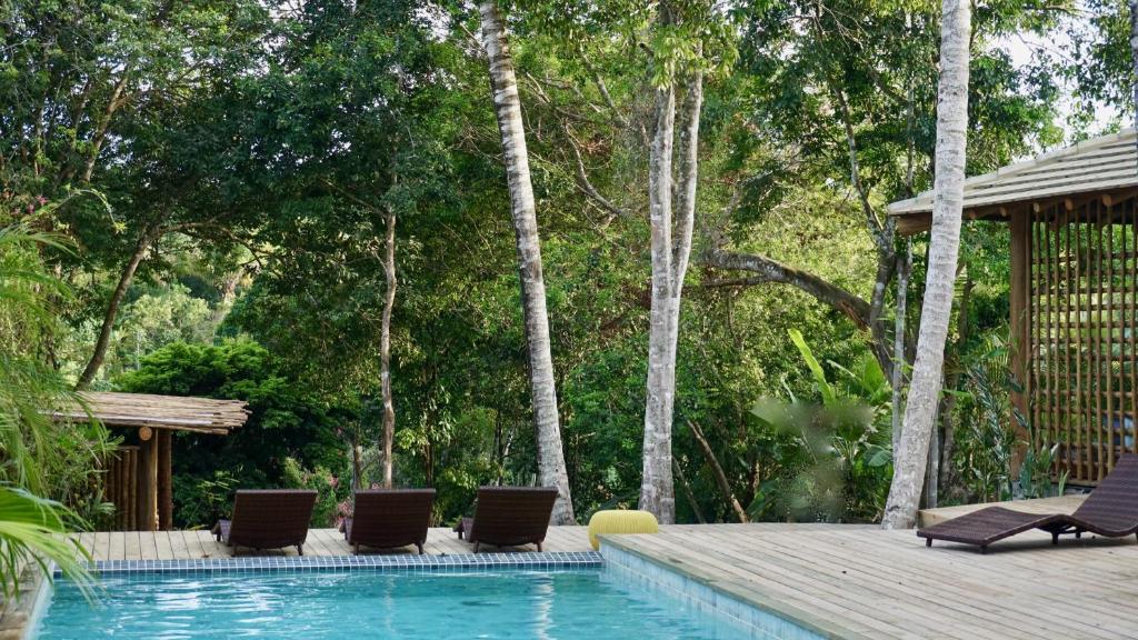 Viagem para Bahia: Piscina de borda infinita em uma pousada tropical, cercada por vegetação exuberante e altas palmeiras. A água azul clara contrasta com as espreguiçadeiras marrons e cadeiras de madeira, criando uma atmosfera serena e convidativa. A estrutura de madeira à esquerda adiciona um toque rústico ao design, enquanto a luz natural filtra-se através das folhas, projetando sombras suaves. A composição harmoniosa entre a arquitetura e a natureza transmite uma sensação de tranquilidade e conexão com a natureza, característica da Pousada Soleluna em Trancoso, Bahia.