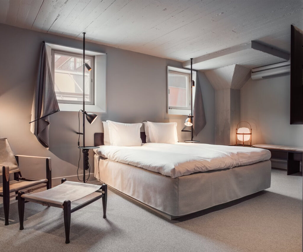 acomodação blique by nobis. moderno, O quarto apresenta uma cama king-size com lençóis brancos impecáveis e travesseiros fofos. Há uma poltrona de madeira e um pufe combinando, além de uma mesa lateral com uma luminária moderna. As paredes são pintadas em tons suaves de cinza, e o teto tem vigas de madeira expostas. Duas janelas com cortinas pretas permitem a entrada de luz natural.