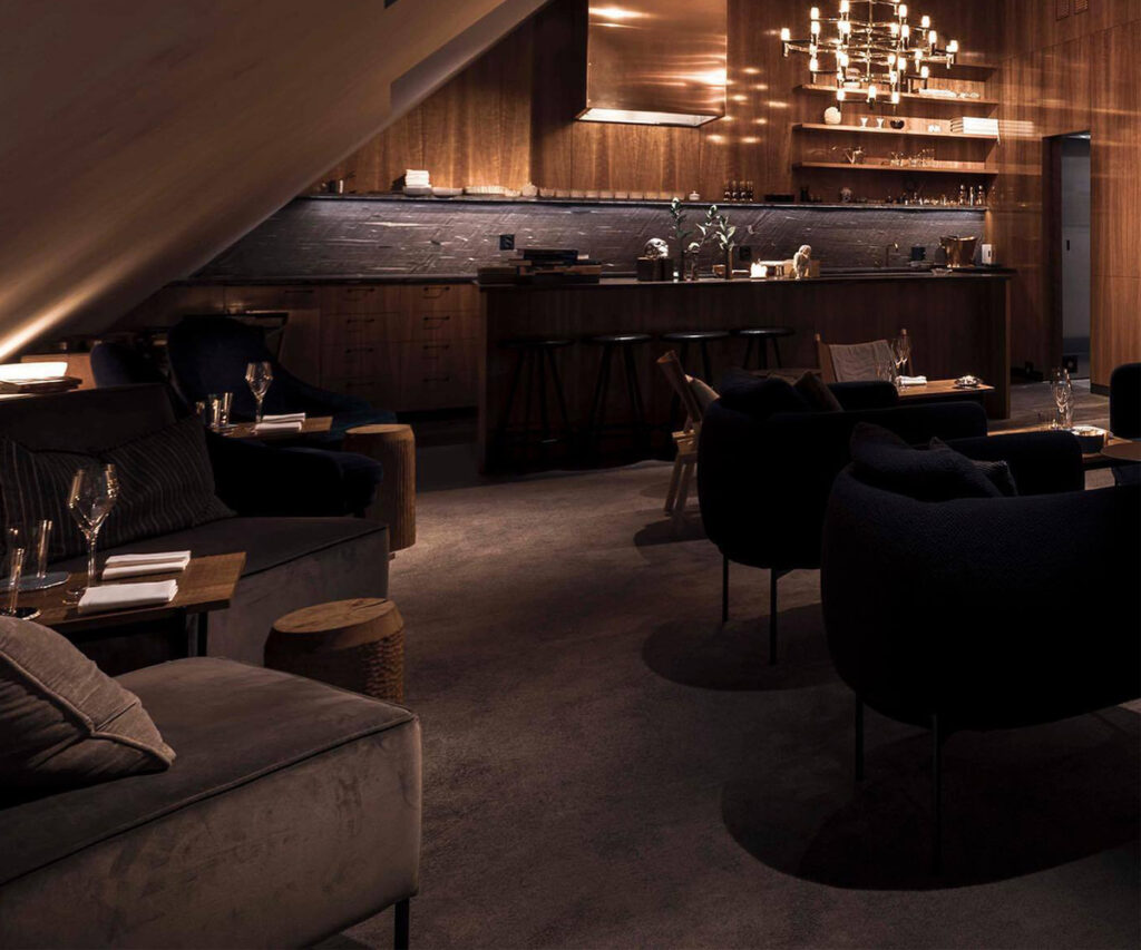 Salão do restaurante Frantzén. A imagem mostra o interior de um salão de restaurante moderno com uma paleta de cores quentes e acolhedoras. O ambiente é caracterizado por móveis de couro escuro, cadeiras confortáveis e uma iluminação suave que cria uma atmosfera íntima. O bar no fundo é equipado com várias garrafas e copos, e há uma grande luminária pendente acima dele. As paredes e o teto são feitos de madeira, adicionando um toque rústico ao design contemporâneo.