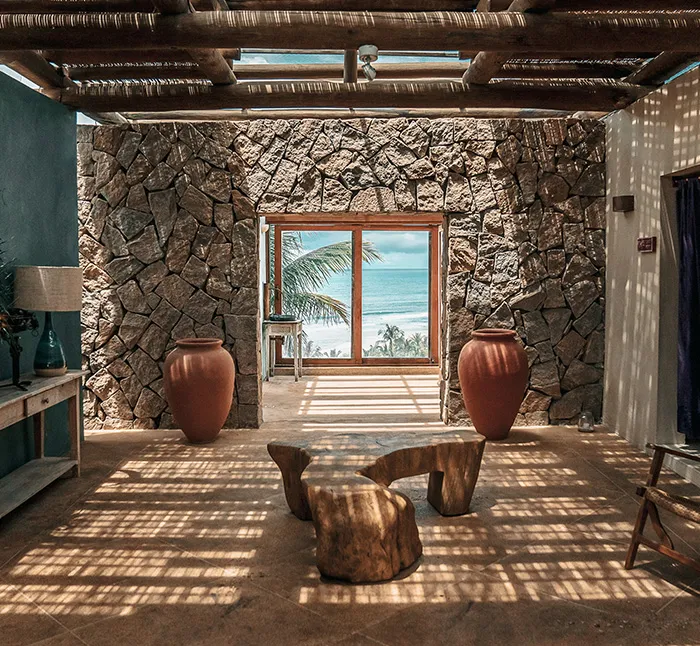 Um interior rústico e acolhedor com paredes de pedra e elementos naturais. A sala é dominada por dois grandes vasos de terracota posicionados de cada lado de uma porta de vidro que revela uma vista deslumbrante do oceano e palmeiras balançando suavemente. A iluminação natural entra por uma claraboia, projetando sombras suaves no chão de concreto. Um banco de madeira esculpida e uma mesa de apoio com uma lâmpada complementam o design minimalista e orgânico. A atmosfera é serena e convidativa, perfeita para uma experiência de viagem tranquila e conectada à natureza, característica do Txai Resort em Trancoso, Bahia.