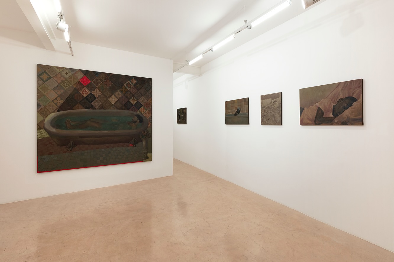 A imagem mostra uma galeria de arte moderna com várias pinturas expostas em uma parede branca. As pinturas retratam cenas de banheiros com elementos surrealistas. O chão é de madeira clara e o teto tem iluminação industrial com tubos de luz. É a Gentil Carioca, uma das principais galerias de arte do rio de janeiro
