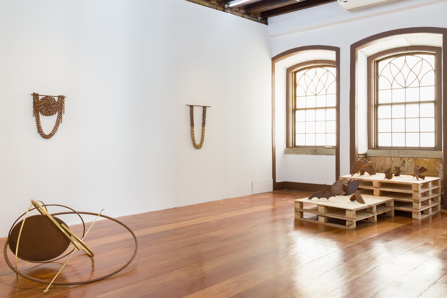 A imagem mostra uma galeria de arte moderna com decoração minimalista e industrial. A sala possui um piso de madeira e paredes brancas. Duas peças de arte estão penduradas na parede, e há uma estrutura circular de metal no chão. No lado direito, há paletes de madeira com objetos sobre eles. As janelas arqueadas com vidros em forma de diamante permitem a entrada de luz natural.