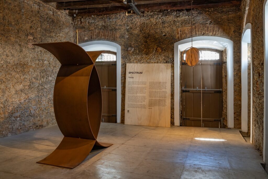 A imagem retrata uma galeria de arte moderna com uma grande escultura de metal oxidado em formato circular. A galeria possui paredes de pedra e um chão de concreto, conferindo um ar industrial ao espaço. Há uma placa informativa de madeira com o título 'Spectrum' e texto descritivo. A iluminação é natural, vinda de janelas arqueadas, e há uma lâmpada pendente em forma de esfera. O ambiente é decorado de forma minimalista, com foco na arte e na simplicidade. É a Gentil Carioca, uma das principais galeras de arte no rio de janeiro
