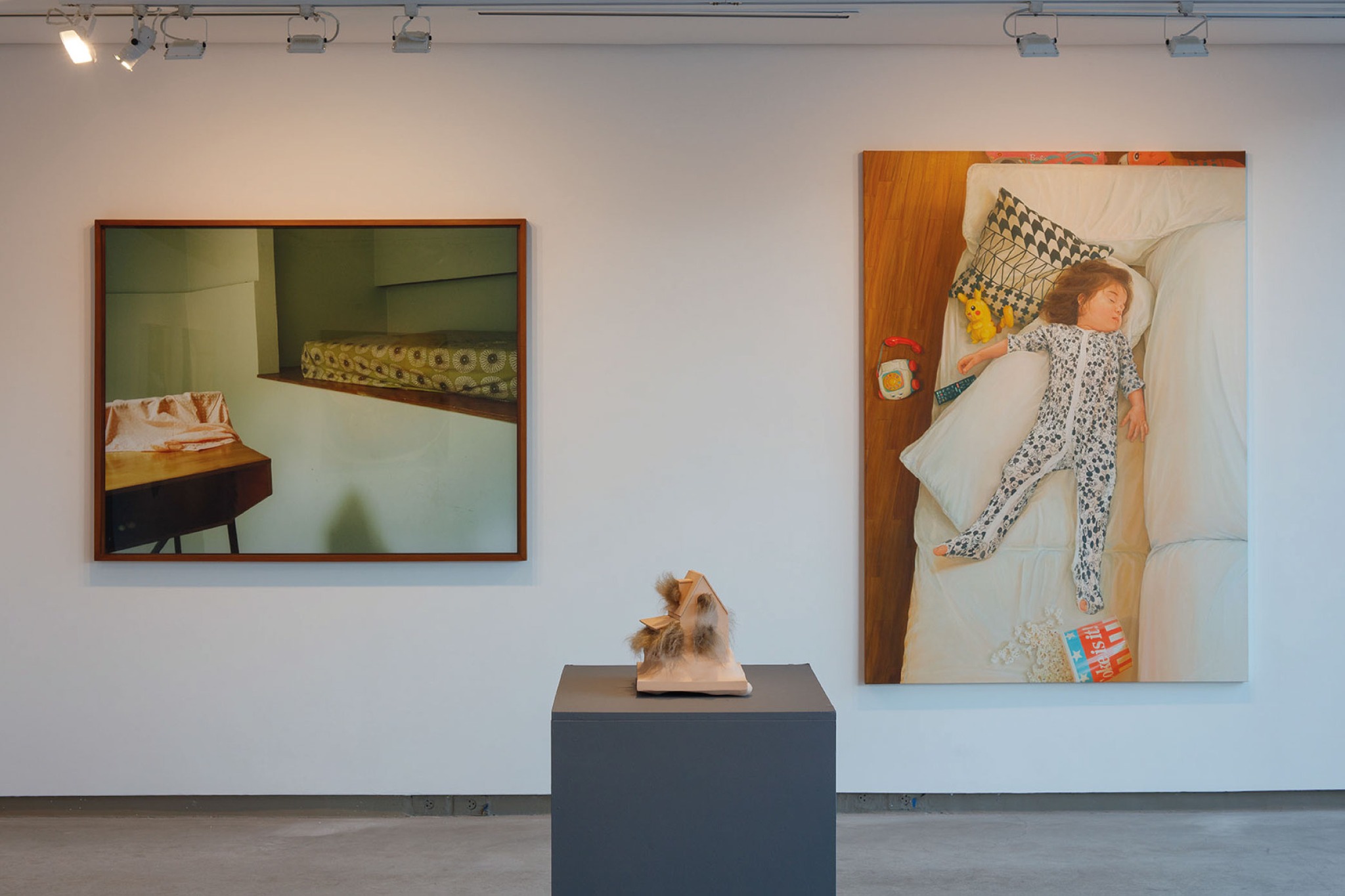 A imagem mostra uma exposição de arte moderna na Anita Schwartz Galeria de Arte no Rio de Janeiro. A sala apresenta várias pinturas coloridas e uma escultura em um pedestal. As paredes são brancas, e o chão é de concreto, proporcionando um fundo neutro para as obras de arte. A iluminação é fornecida por luzes de trilho no teto, destacando cada peça.