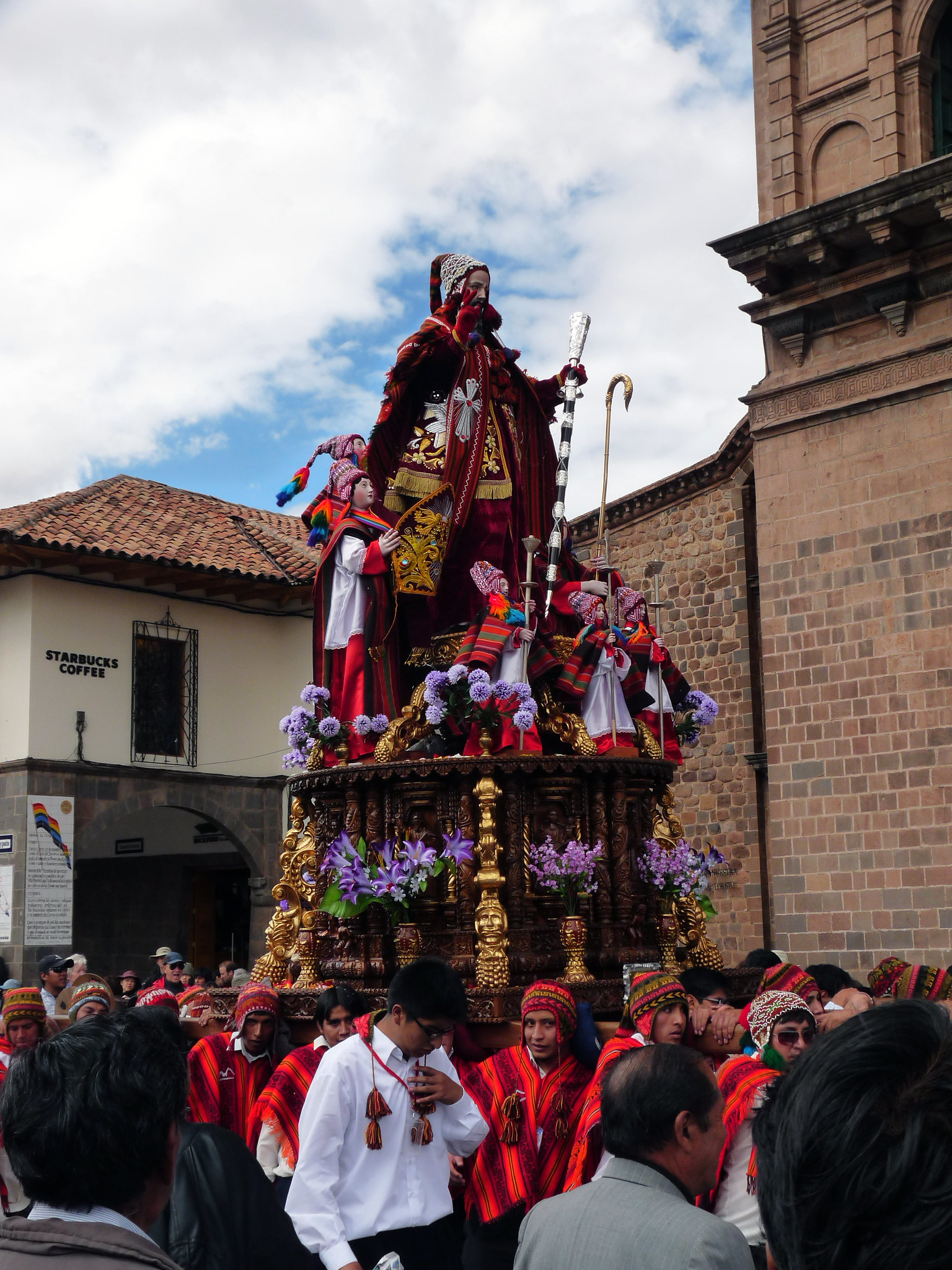Dicas de viagem pelo Peru: A imagem captura uma vibrante procissão de Corpus Christi em Cusco, Peru. A procissão é liderada por uma estátua ornamentada de uma figura religiosa, adornada com tecidos vermelhos e dourados, e cercada por outras estátuas menores. Os participantes, vestidos com trajes tradicionais, carregam a estátua em uma plataforma dourada decorada com flores roxas. O fundo inclui um edifício de tijolos e uma placa de uma cafeteria Starbucks.