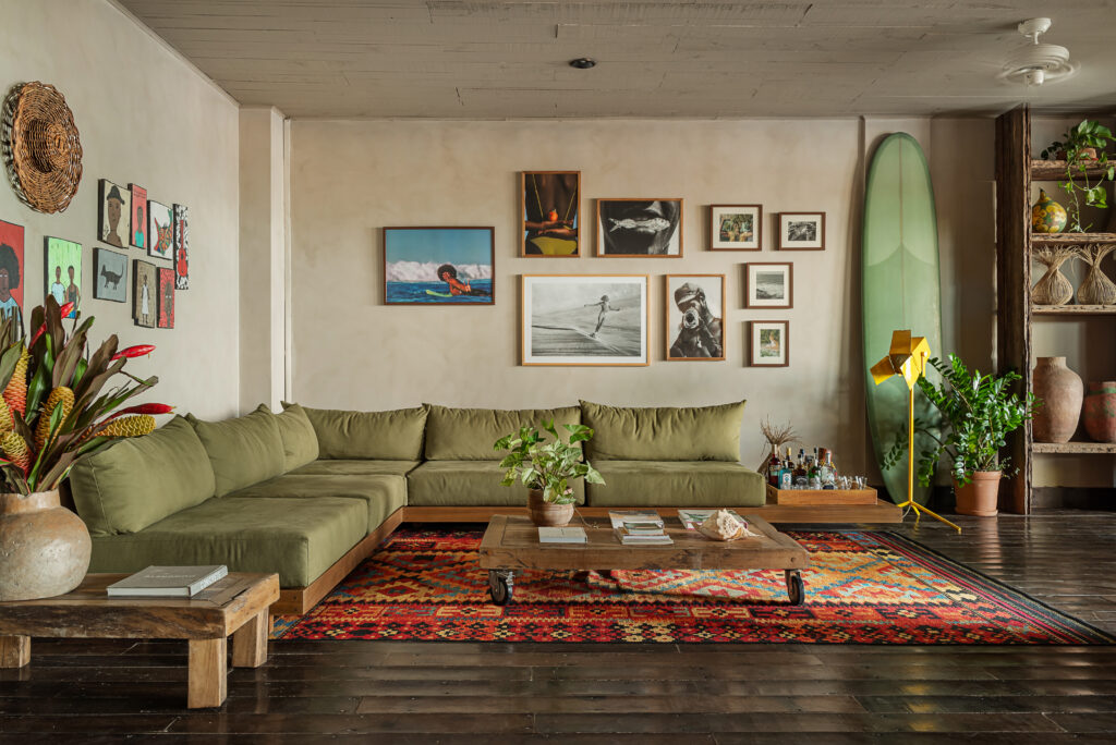 Sala de estar moderna com sofá verde vibrante e tapete colorido. A parede é adornada com uma variedade de quadros e fotos emolduradas, incluindo imagens de praia e surfistas, criando uma atmosfera descontraída e artística. Uma prancha de surfe verde se destaca ao lado de prateleiras cheias de plantas e vasos. A decoração inclui vasos grandes com plantas exuberantes e uma mesa de centro de madeira com livros e objetos decorativos. A iluminação natural e as cores quentes transmitem uma sensação de relaxamento e conexão com a natureza, típica de uma viagem para a Bahia.