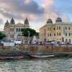 Praça do Marco Zero em Recife, Pernambuco, Brasil. A imagem captura uma cena vibrante ao pôr do sol com edifícios coloniais de estilo arquitetônico português, predominantemente amarelos e brancos, com detalhes em tons terrosos. Os edifícios apresentam cúpulas e janelas arqueadas, conferindo um ar histórico à paisagem. Em primeiro plano, barcos tradicionais vermelhos e brancos flutuam na água calma, adicionando um toque de charme marítimo. Pessoas estão reunidas ao longo da orla, desfrutando da vista e do ambiente relaxado. A composição é harmoniosa, com o céu nublado e a iluminação suave criando uma atmosfera tranquila e convidativa. O que fazer emRecife.