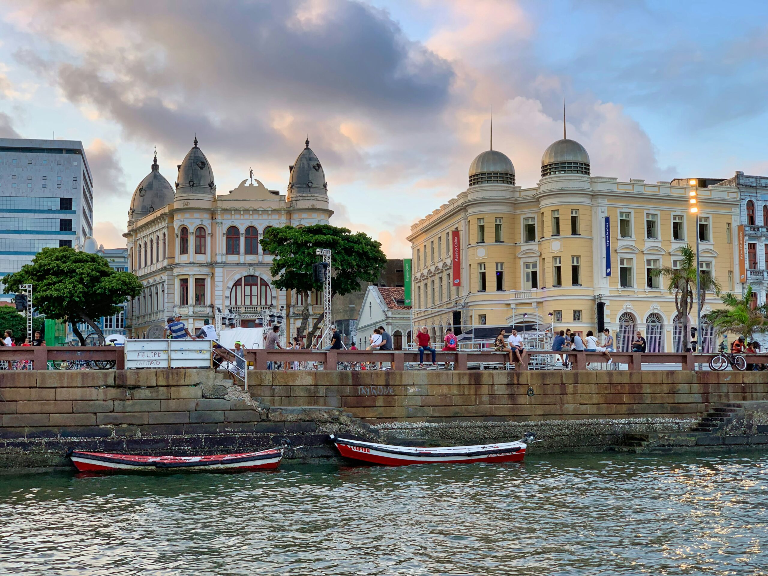 Praça do Marco Zero em Recife, Pernambuco, Brasil. A imagem captura uma cena vibrante ao pôr do sol com edifícios coloniais de estilo arquitetônico português, predominantemente amarelos e brancos, com detalhes em tons terrosos. Os edifícios apresentam cúpulas e janelas arqueadas, conferindo um ar histórico à paisagem. Em primeiro plano, barcos tradicionais vermelhos e brancos flutuam na água calma, adicionando um toque de charme marítimo. Pessoas estão reunidas ao longo da orla, desfrutando da vista e do ambiente relaxado. A composição é harmoniosa, com o céu nublado e a iluminação suave criando uma atmosfera tranquila e convidativa. O que fazer emRecife.