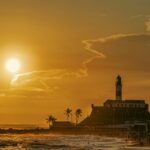 Pôr do sol vibrante na praia com um farol icônico ao fundo. O céu é pintado em tons de laranja e amarelo, com nuvens esvoaçantes adicionando textura. O farol, alto e com listras contrastantes, destaca-se contra a silhueta do horizonte. Palmeiras balançam suavemente na praia arenosa, onde pessoas aproveitam o mar. A cena transmite uma atmosfera serena e convidativa, típica de uma viagem à Bahia, com um design harmonioso que combina a beleza natural com a arquitetura histórica.