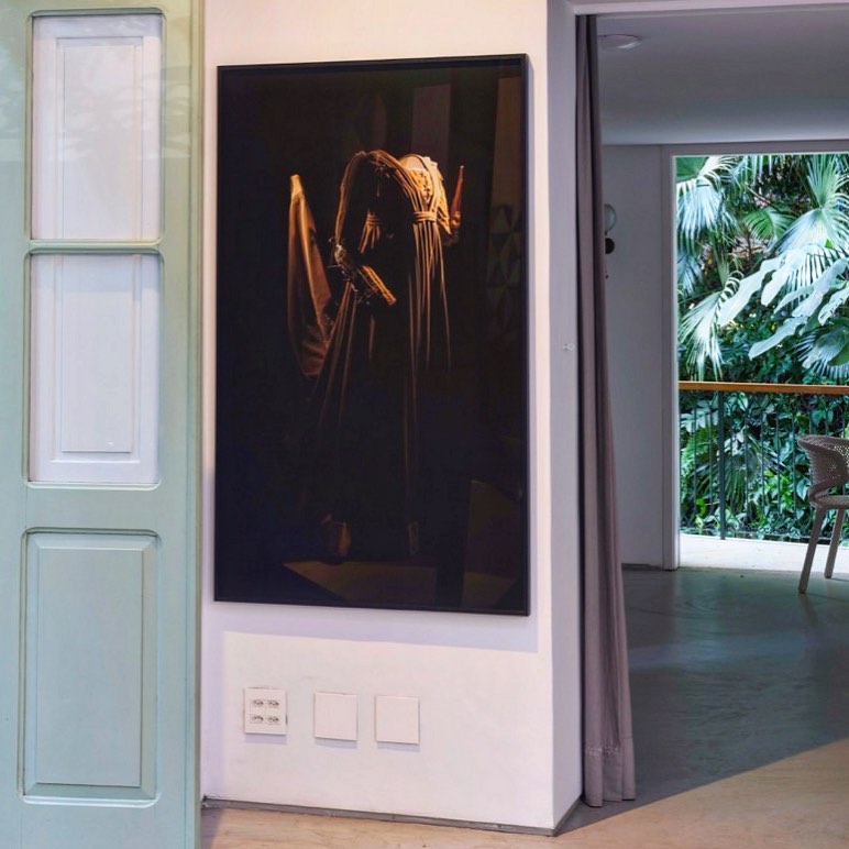 A imagem mostra uma exposição de um vestido usado pela atriz Olívia Hussey no filme 'Romeu e Julieta', de Franco Zeffirelli. O vestido está exibido em um manequim dentro de uma vitrine com fundo preto, destacando-se contra uma parede branca. A exposição está localizada na Galeria da Gávea no Rio de Janeiro. A decoração do ambiente é minimalista, com uma paleta de cores que inclui branco e tons neutros. A vitrine é cercada por uma moldura branca simples, e há uma porta verde à esquerda com painéis de vidro. O ambiente parece ser uma galeria de arte moderna, com elementos naturais visíveis através de uma porta aberta que leva a um pátio externo com plantas tropicais.