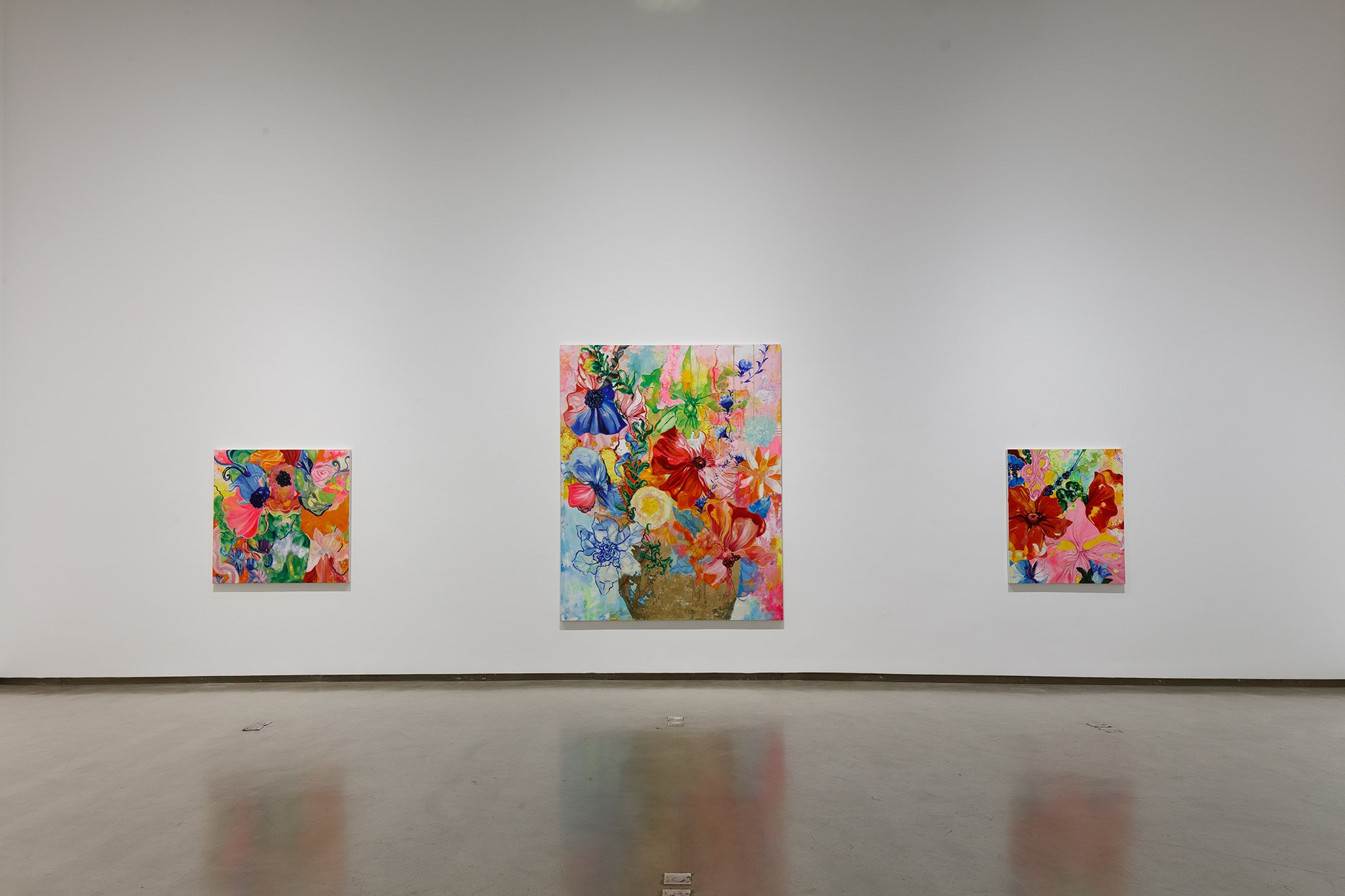 A imagem mostra uma exposição de arte na Anita Schwartz Galeria de Arte no Rio de Janeiro. A exposição apresenta três pinturas vibrantes de flores em uma parede branca. As pinturas são coloridas e detalhadas, exibindo uma variedade de flores e folhas. O ambiente é minimalista, com um chão de concreto polido e iluminação natural.