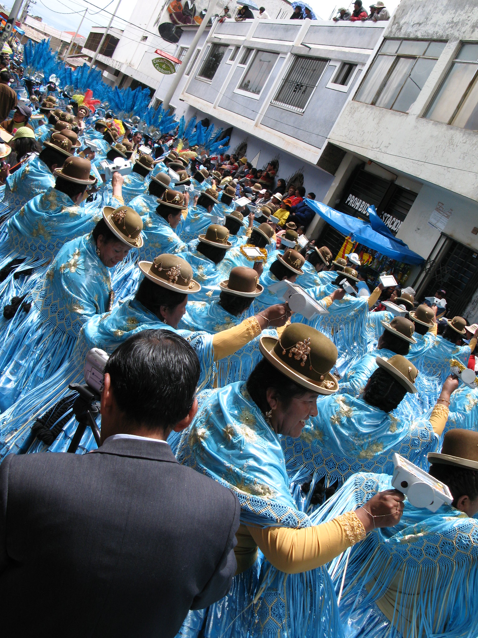 A imagem captura um vibrante desfile festivo em Puno, Peru, com participantes vestidos com trajes tradicionais azuis e chapéus. O evento faz parte do Festival da Virgen de Candelaria, celebrado anualmente. A multidão está reunida em uma rua movimentada, com espectadores assistindo de janelas e varandas. Os trajes são adornados com detalhes dourados e os participantes estão segurando objetos brancos, possivelmente parte da cerimônia.