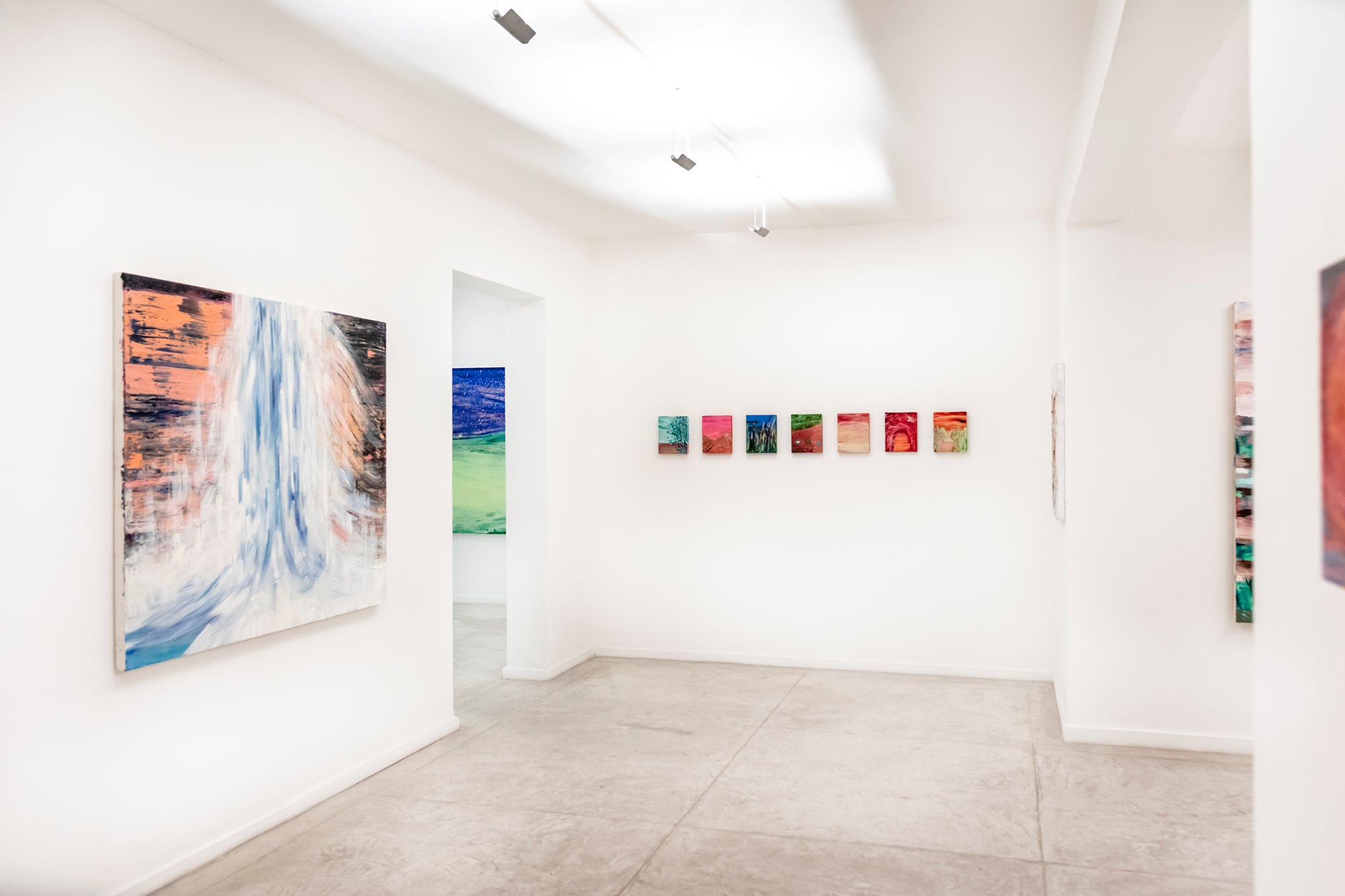 A imagem mostra a interior da Galeria Mercedes Viegas no Rio de Janeiro. A sala é ampla e bem iluminada, com paredes e teto brancos. As principais características são as várias pinturas expostas nas paredes. À esquerda, há uma grande pintura abstrata com tons de azul, branco e laranja. No centro, uma fileira de pinturas menores com cores vibrantes, incluindo vermelho, verde e azul. À direita, uma pintura vertical com tons de vermelho e verde. O chão é de concreto, e a iluminação é natural, criando um ambiente minimalista e moderno.