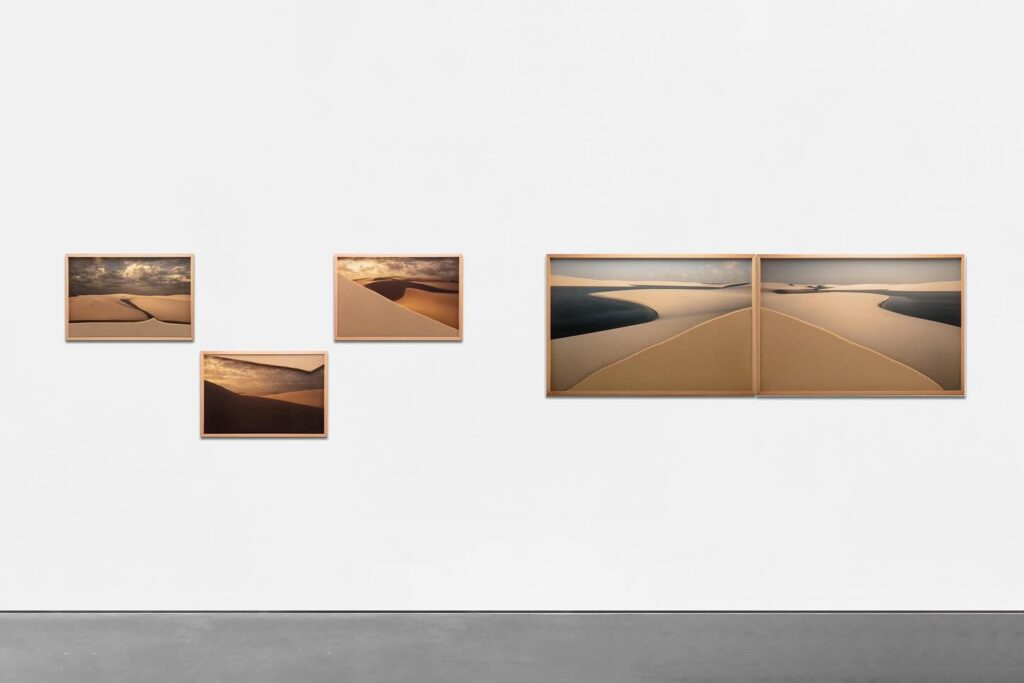 A imagem mostra uma exposição de arte na Galeria de Arte Silvia Cintra, localizada no Rio de Janeiro. A exposição apresenta quatro fotografias emolduradas de dunas de areia, dispostas de forma organizada em uma parede branca. As fotografias capturam a beleza natural das dunas sob diferentes condições de luz, destacando as formas e texturas da paisagem. A galeria possui um design minimalista com um foco na simplicidade e na valorização das obras de arte.