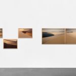 A imagem mostra uma exposição de arte na Galeria de Arte Silvia Cintra, localizada no Rio de Janeiro. A exposição apresenta quatro fotografias emolduradas de dunas de areia, dispostas de forma organizada em uma parede branca. As fotografias capturam a beleza natural das dunas sob diferentes condições de luz, destacando as formas e texturas da paisagem. A galeria possui um design minimalista com um foco na simplicidade e na valorização das obras de arte.