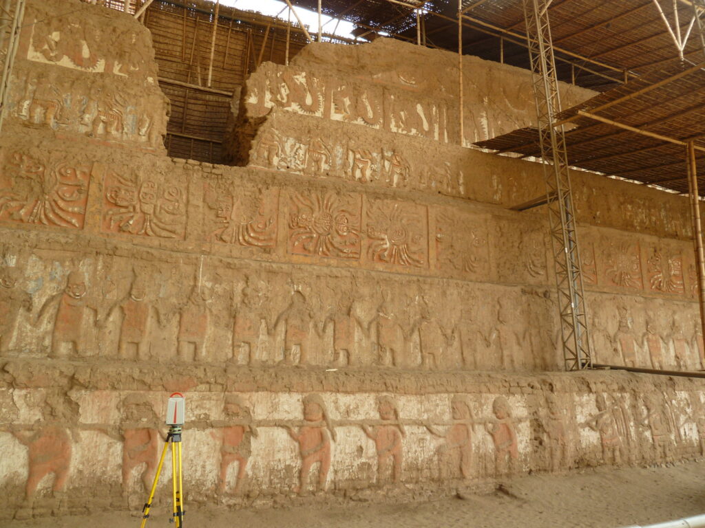 Dicas de viagem pelo Peru: A imagem mostra uma estrutura arqueológica com entalhes detalhados em uma parede de pedra. O local parece ser um templo, possivelmente um dos templos de Huacas del Sol e la Luna perto de Trujillo. Equipamentos de medição estão presentes, indicando trabalho de preservação ou restauração. A estrutura é coberta por um telhado de madeira e há andaimes ao redor.