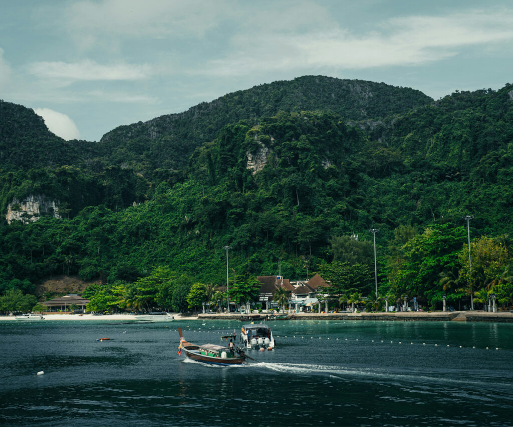 Praia das ilhas phi phi com montanhas ao fundo e barcos no mar. Barcos navegando em águas azuis-esverdeadas em frente a uma praia tropical. Ao fundo, montanhas exuberantes e verdes com vegetação densa. A cena é serena, com um clima tropical e uma atmosfera relaxante. Estruturas de madeira e palmeiras ao longo da costa, sugerindo um ambiente de resort ou vila de praia. A composição equilibra os elementos naturais com toques de design rústico, criando uma experiência visual harmoniosa e convidativa.