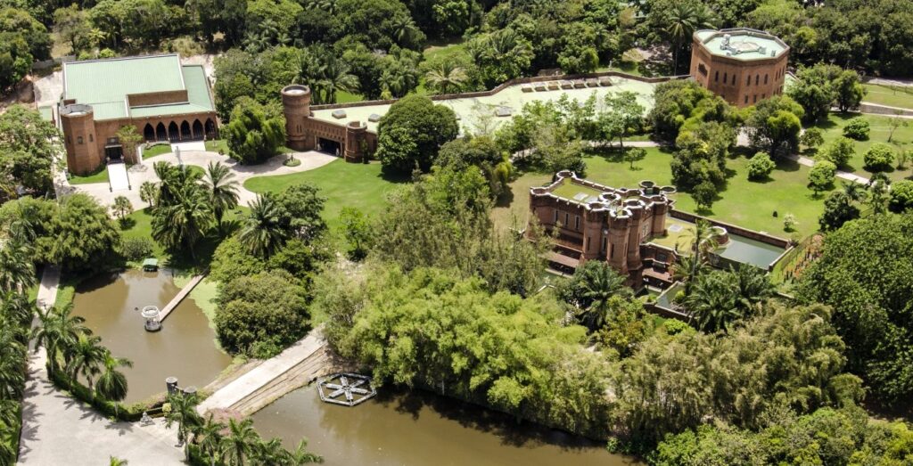 O que fazer em Recife: A imagem apresenta uma vista aérea do Instituto Brennand, um complexo arquitetônico impressionante em Recife. O edifício principal, de estilo Tudor, destaca-se com suas paredes de tijolo vermelho e telhado verde, reminiscente da arquitetura gótica medieval. Ao redor, extensos jardins exuberantes e palmeiras pontuam a paisagem, criando uma atmosfera serena e convidativa. Um lago com uma pequena ilha e uma ponte de madeira adiciona um toque de tranquilidade à cena. A composição harmoniosa de estruturas históricas e natureza exuberante destaca a curadoria cuidadosa do acervo e a estética do local.