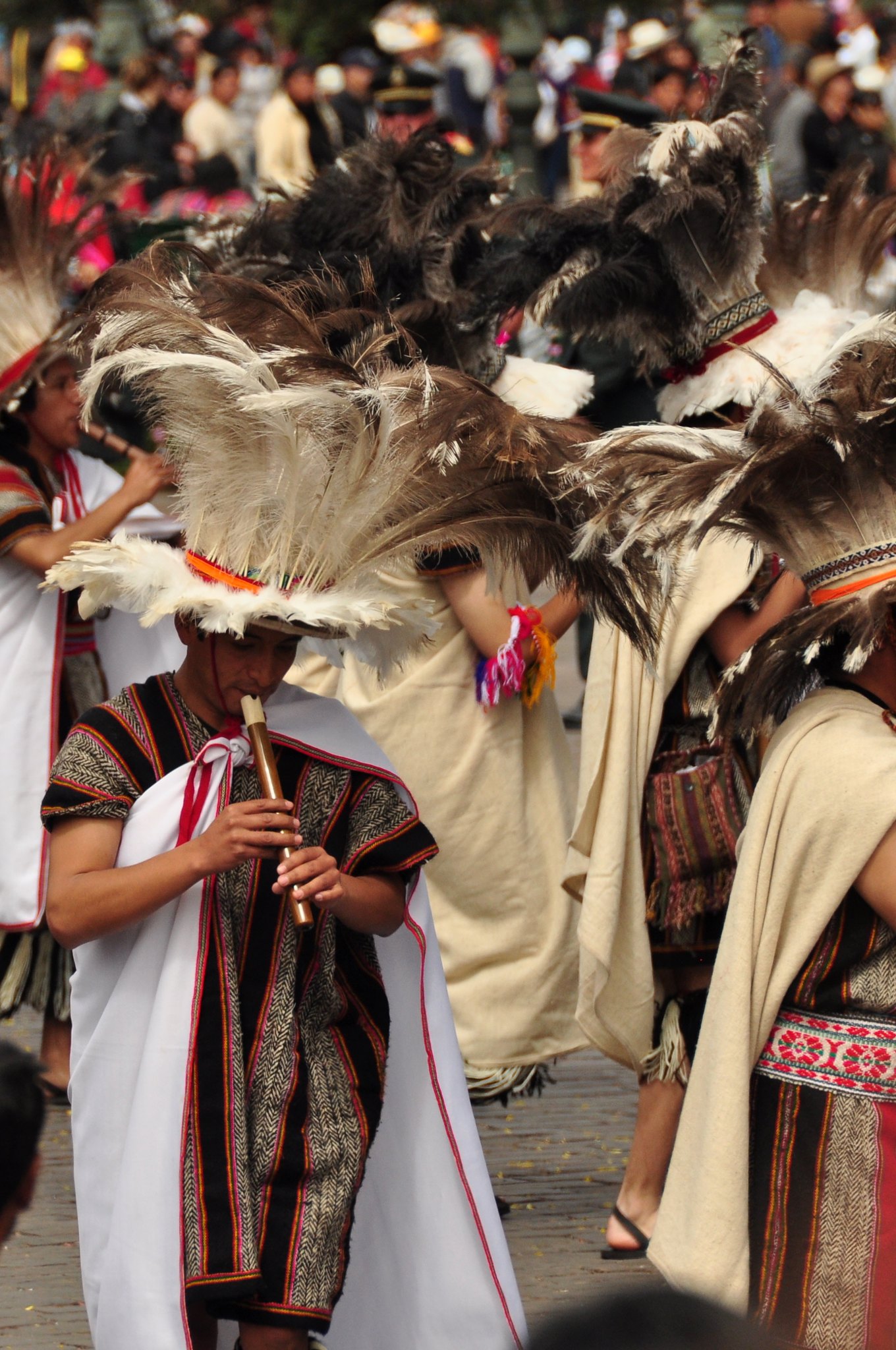 Dicas de viagem pelo Peru: A imagem captura uma cena vibrante da celebração do Inti Raymi em Cusco, Peru. Os participantes estão adornados com trajes tradicionais, incluindo grandes adereços de penas e roupas coloridas. A paleta de cores predominantes inclui tons de vermelho, branco e preto, com detalhes em dourado e laranja. Os materiais usados incluem tecidos, penas e acessórios de metal. O estilo de decoração é cultural e tradicional, refletindo a herança inca. O tipo de ambiente é uma celebração pública ao ar livre, com um fundo de multidão e elementos naturais.
