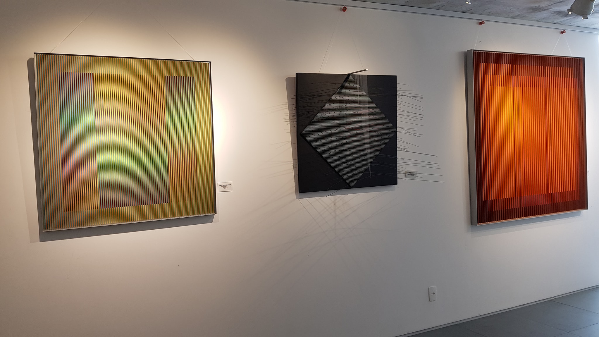 A imagem mostra uma exposição de arte cinética na galeria de Ipanema, no Rio de Janeiro. As obras de Cruz Diez e Jesus Soto estão expostas lado a lado, apresentando uma paleta de cores vibrantes e geometrias abstratas. As peças são predominantemente coloridas em tons de amarelo, preto e laranja, com linhas verticais e formas geométricas que criam um efeito visual dinâmico. O ambiente é moderno e minimalista, com paredes brancas e iluminação cuidadosa que realça as obras de arte.