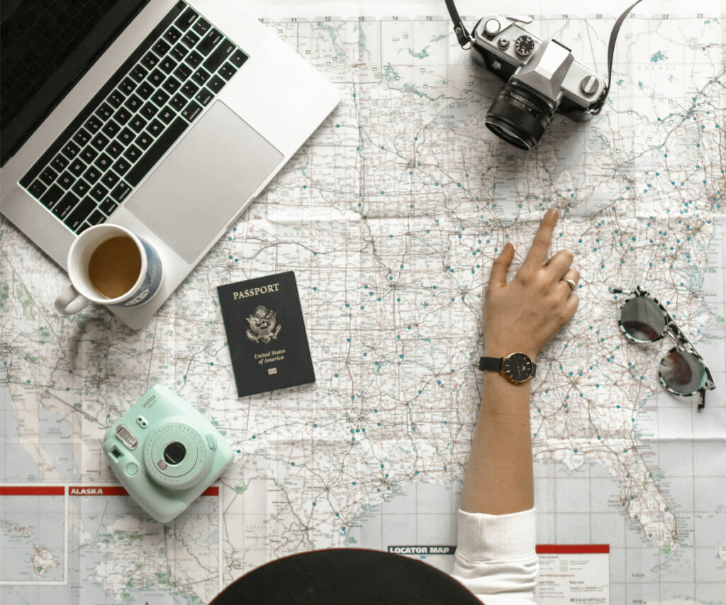 pessoa analisando um mapa colocado em cima de uma mesa junto com um notebook, duas câmeras fotográficas, um passafprte, uma caneca e um óculos escuros para fazer um roteiro de viagem
